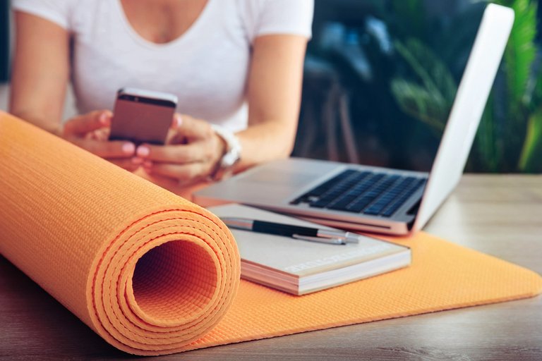 A half-rolled orange yoga mat on a table with a book, a pen, and a laptop, with a woman holding a cell phone in the background 